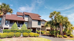 A multi-story hotel with a symmetrical facade, a central entrance, and a well-maintained garden with various shrubs and palm trees. - Lakeside Updated Villa: Pool and Beach Access (Pawleys Island)