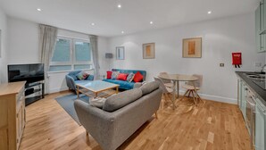 Un salon moderne avec un canapé gris, un canapé bleu avec des coussins rouges, une table basse en bois, une table à manger en verre avec des chaises, un téléviseur et un coin cuisine avec un évier et des placards.
