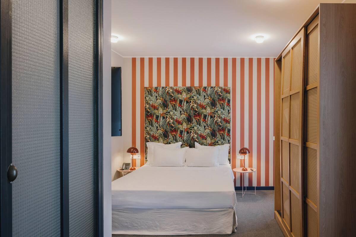 A hotel room with a large bed, striped wallpaper, a tropical-themed headboard, and a wooden wardrobe.