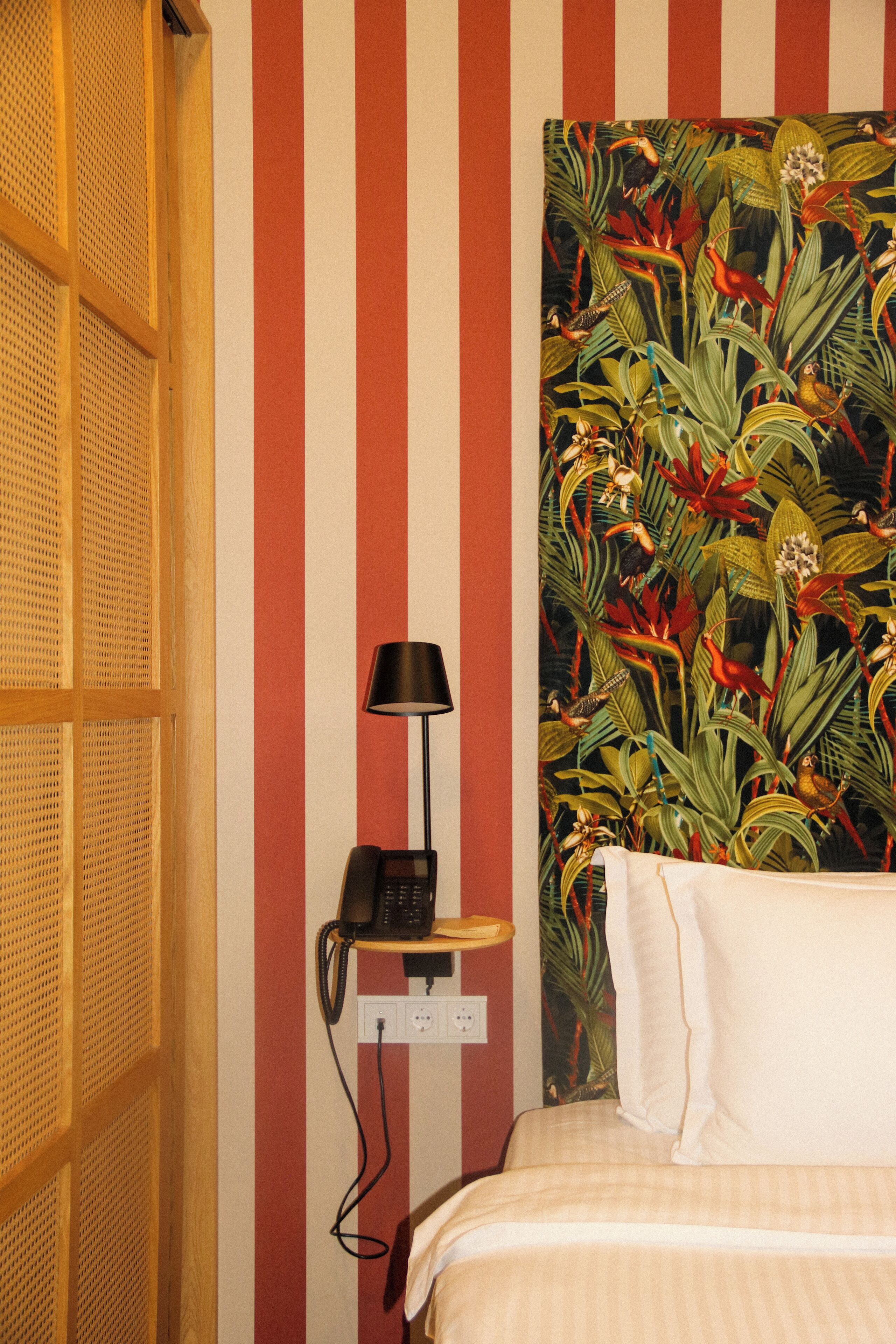 A room with a bed, a telephone on a small table, and a wall with vertical stripes and a tropical plant mural.