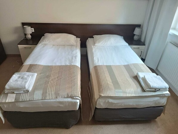 A hotel room with two single beds, each with a pillow and a folded towel. There is a nightstand with a lamp and a phone on the left side.
