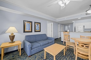 A hotel room with a blue sofa, a wooden table, a lamp, and a kitchenette area.