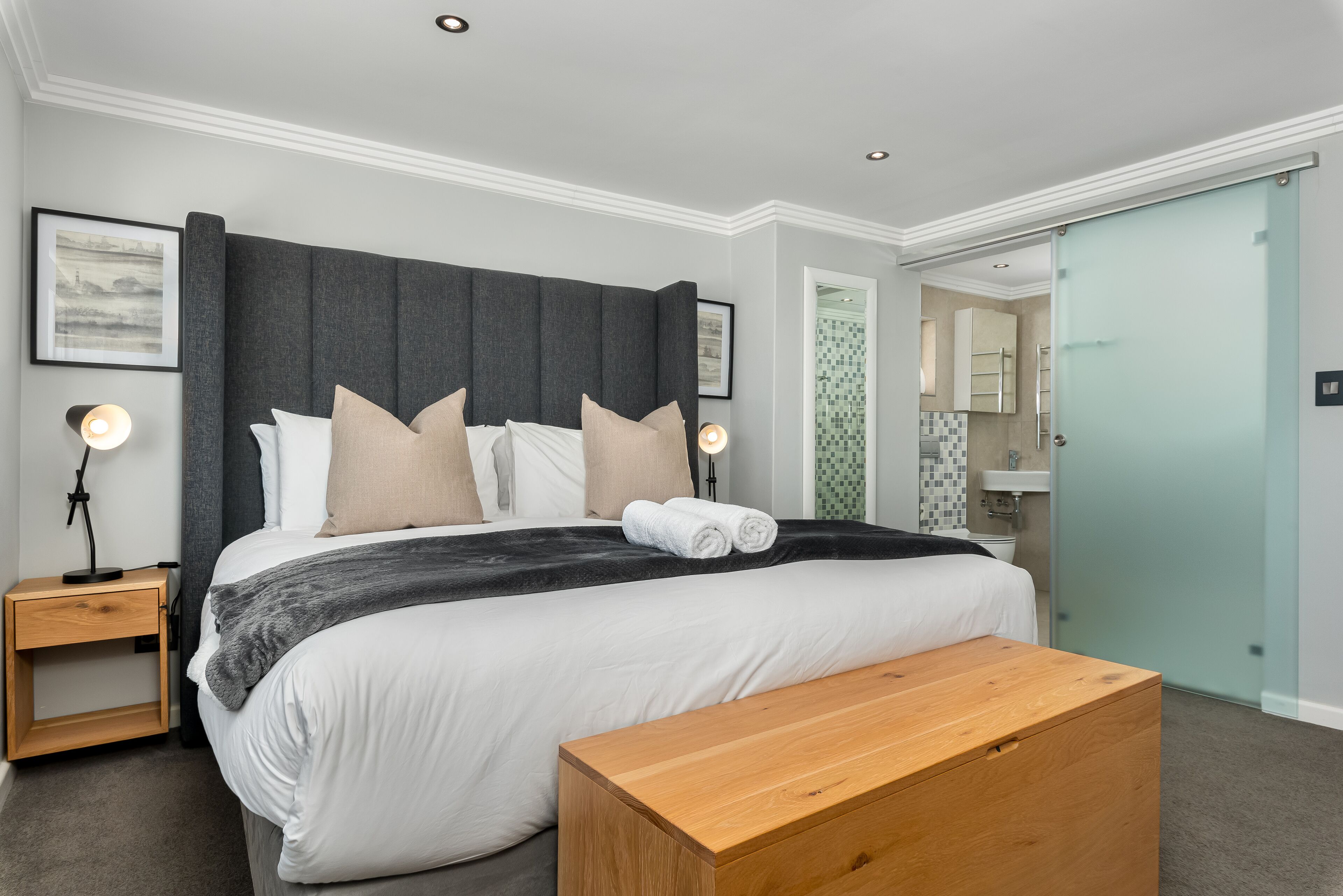 A modern bedroom with a large bed, bedside tables, a wooden bench, and a bathroom visible through a glass door.