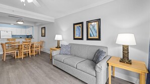A living room with a grey sofa, wooden dining table, and chairs, a ceiling fan, and framed artwork on the wall.
