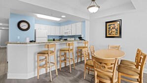 A modern kitchen with white cabinets, a central island with bar stools, a dining table with chairs, a clock on the wall, and a framed picture.