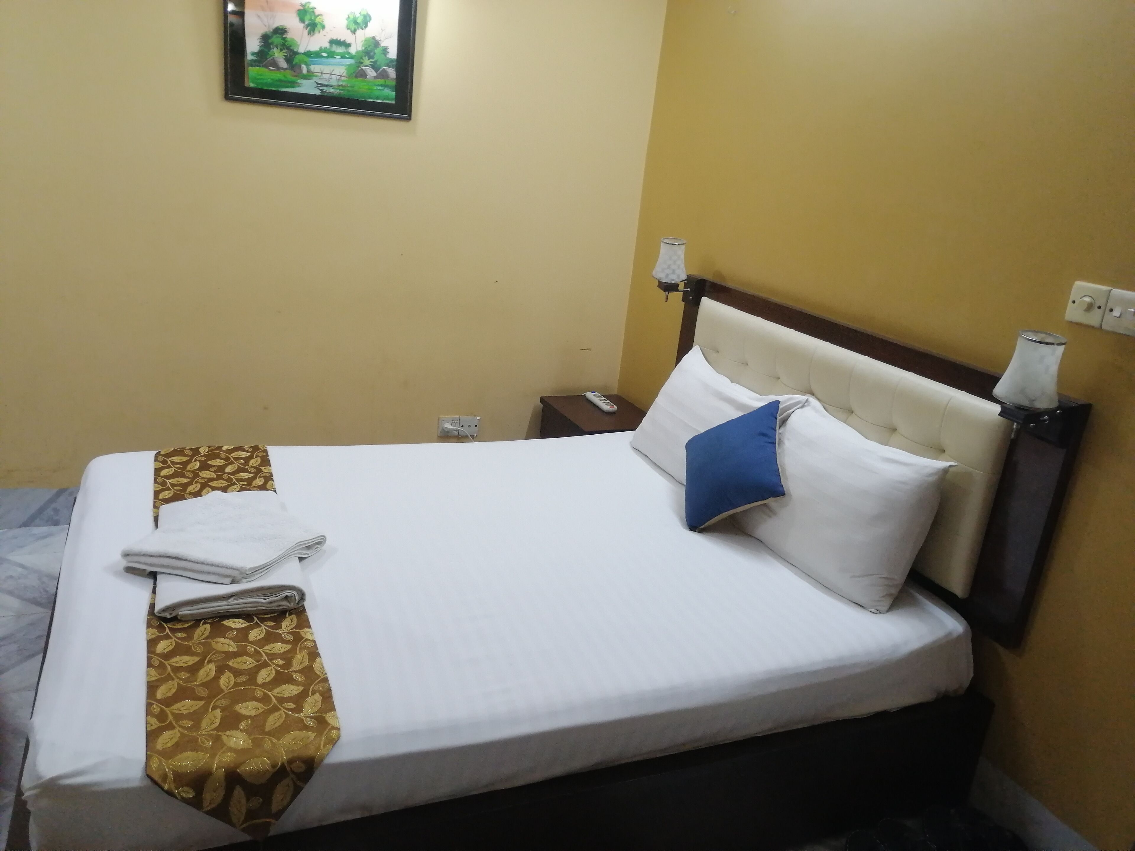 A neatly made bed with white linens, a blue pillow, and a brown patterned throw. There is a remote control on a nightstand beside the bed.