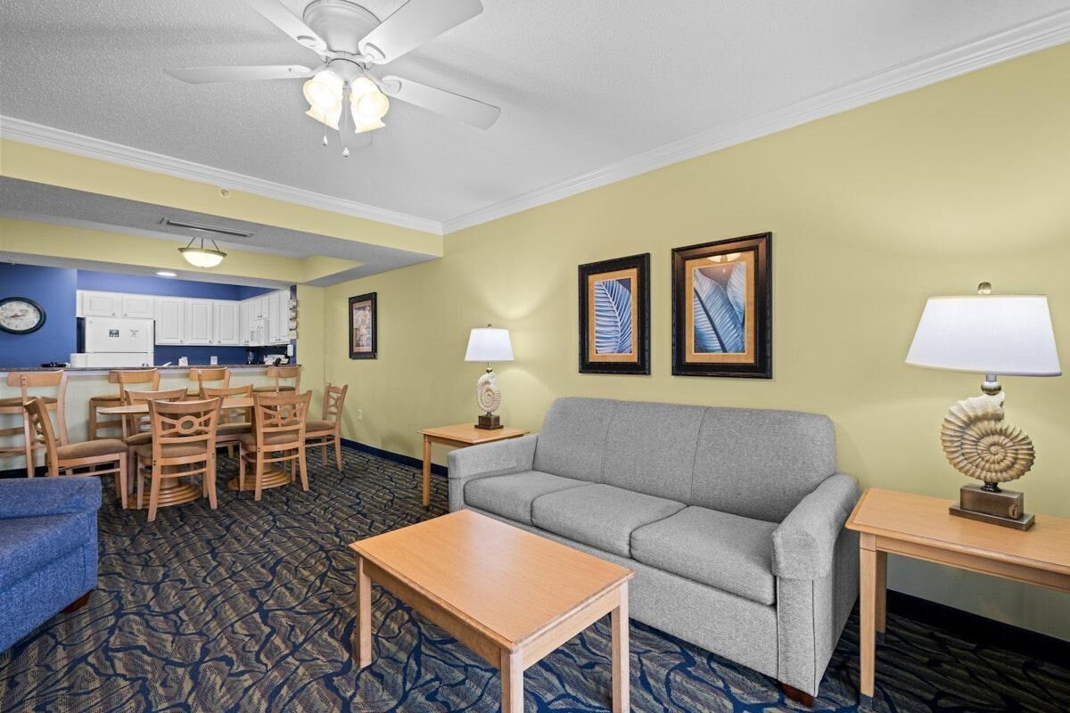 Une chambre d’hôtel équipée d’un canapé, d’une table à manger, de chaises, d’une petite table avec une lampe, d’un ventilateur de plafond et d’œuvres d’art encadrées accrochées au mur.