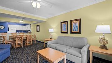 Une chambre d’hôtel équipée d’un canapé, d’une table à manger, de chaises, d’une petite table avec une lampe, d’un ventilateur de plafond et d’œuvres d’art encadrées accrochées au mur.