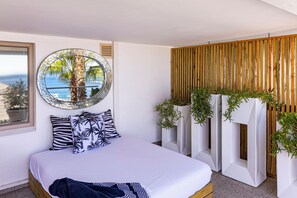 Una camera da letto con un letto, uno specchio rotondo, una finestra vista mare e un divisorio in legno con piante.