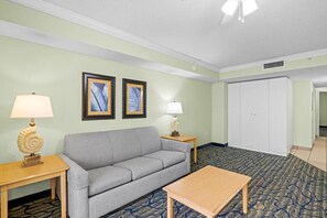 A hotel room with a grey sofa, two side tables with lamps, a wooden coffee table, a patterned carpet, and a wardrobe. - Unwind 1BR Oceanfront Queen Deluxe Condo 4TH Floor (Myrtle Beach)