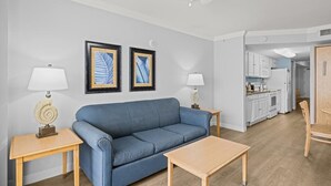 A living room with a blue sofa, two side tables with lamps, framed artwork on the wall, and a view into the kitchen. - Coastal Elegance OF King Suite Escape 4th Floor (Myrtle Beach)