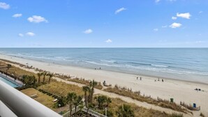 Uitzicht op een strand vanaf een balkon, met heldere lucht, een zandstrand en enkele mensen die wandelen.