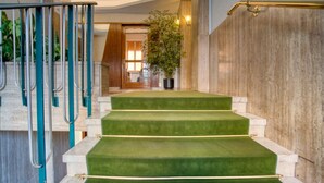 Un escalier dont les marches sont recouvertes d’une moquette verte, avec des rampes en métal et un pot de fleurs placé en haut.