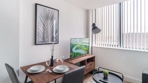 Un comedor moderno con una mesa de madera preparada para dos personas, un televisor de pantalla plana y un ventanal con persianas verticales.