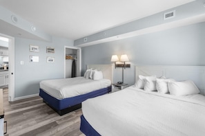 Habitación de hotel con dos camas, un armario, una mesita de noche con lámparas y una kitchenette al fondo.
