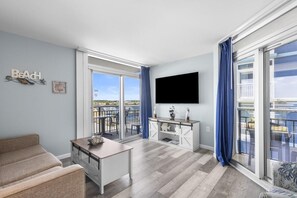 A modern living room with a flat-screen TV, a sofa, a dining table, and a balcony with a view.