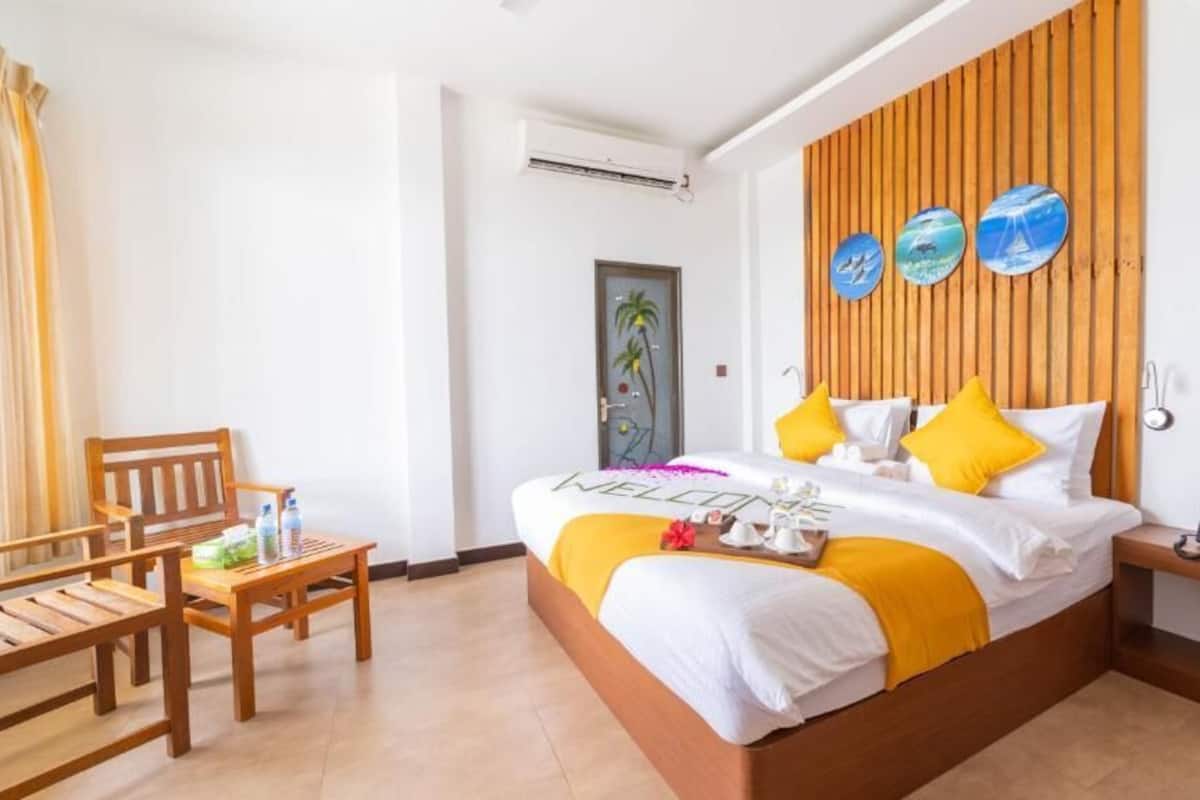 A hotel room with a wooden bed, a wooden side table with a phone, a wooden chair, and a small table with a bottle and a glass.