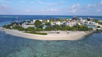 An aerial view of a coastal resort with a sandy beach, clear turquoise waters, and a variety of buildings including a prominent white structure with a red roof.