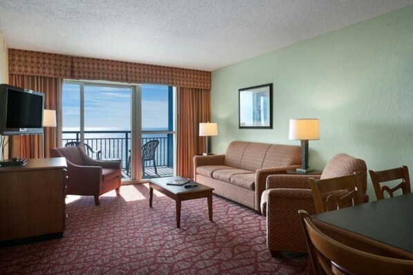 A hotel room with a balcony, a sofa, a desk, a television, and two lamps. - Attractive Oceanfront Condo at Bay View Resort (Myrtle Beach)