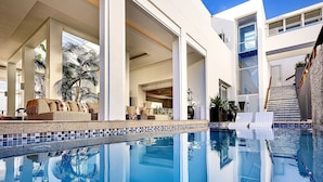 Un área de piscina moderna con agua azul cristalina, un espacio de descanso con sofás y una escalera que conduce a un nivel superior.