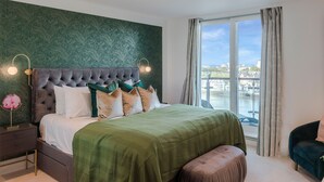 Een moderne slaapkamer met een groot bed, groen beddengoed, een gestoffeerd hoofdeinde, een nachtkastje met een lamp en een uitzicht op een stadsbeeld door een groot raam.