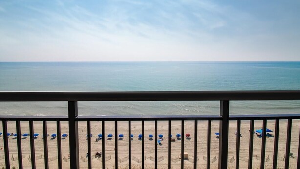 A beach view from a balcony with a railing, featuring a sandy beach with umbrellas and a clear sky. - Wave Watcher's Haven: 2BR 2 Baths, Oceanfront (Myrtle Beach)