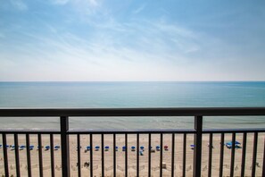 Blick auf einen Strand vom Balkon mit Geländer. Man sieht einen Sandstrand mit Sonnenschirmen und einen klaren Himmel.