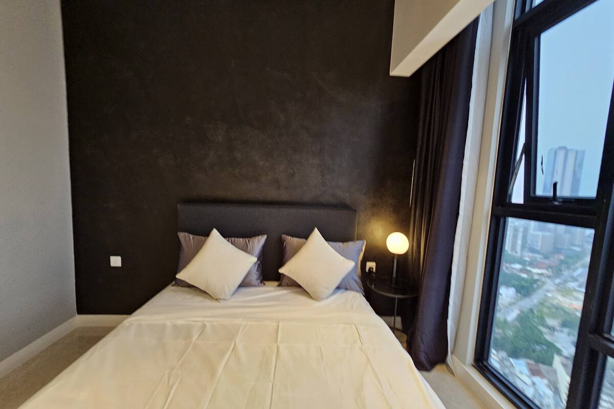 A modern hotel room with a large bed, dark walls, and a window offering a city view.