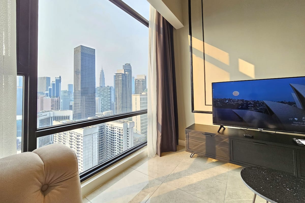 A modern hotel room with a large window offering a city skyline view, a comfortable sofa, a flat-screen TV, and a minimalist console table.