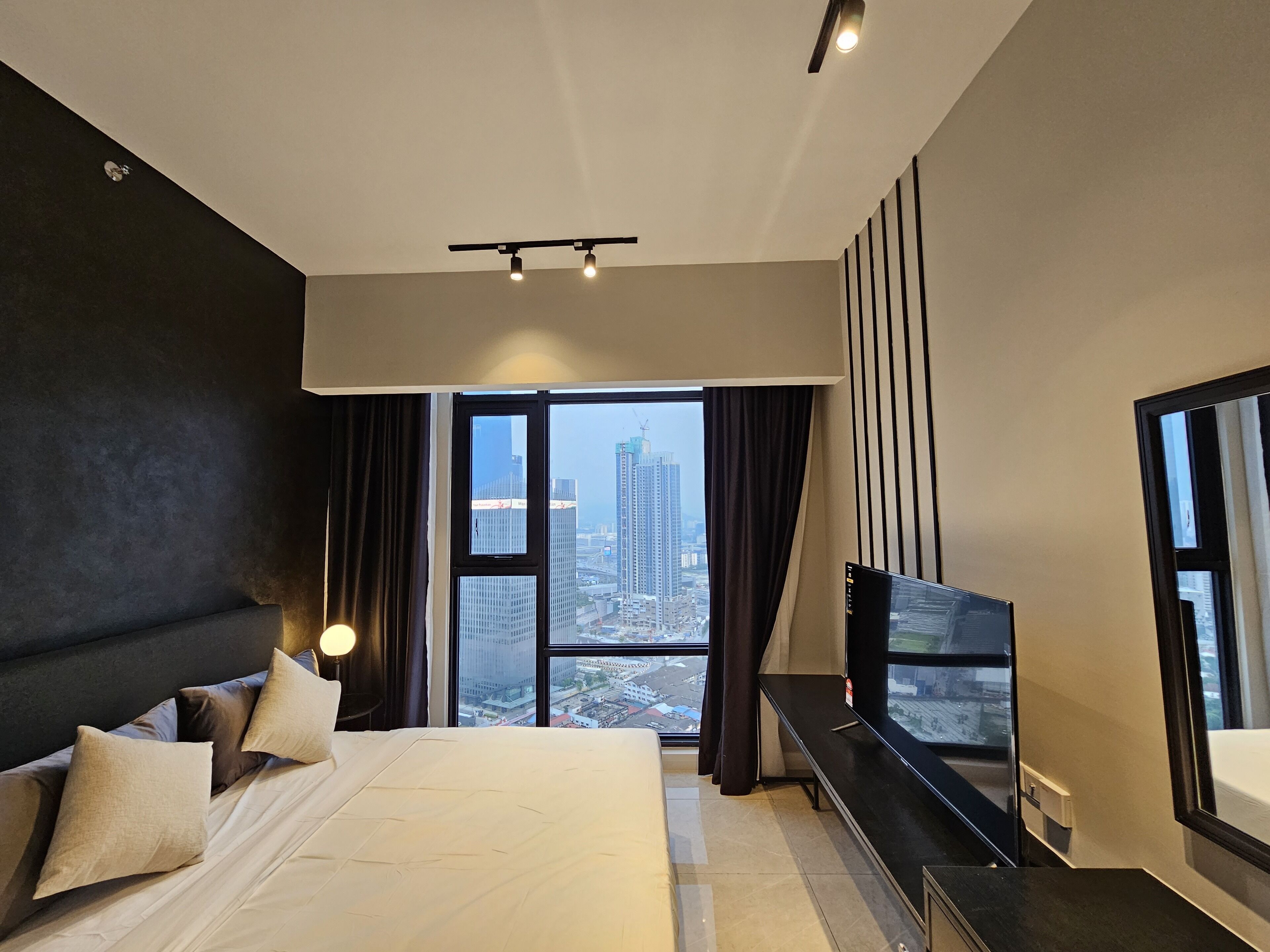A modern hotel room with a large bed, a flat-screen TV, and a city view through large windows.