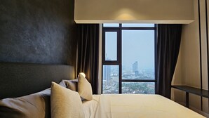 A modern hotel room with a large bed, dark walls, and a view of a cityscape through large windows.