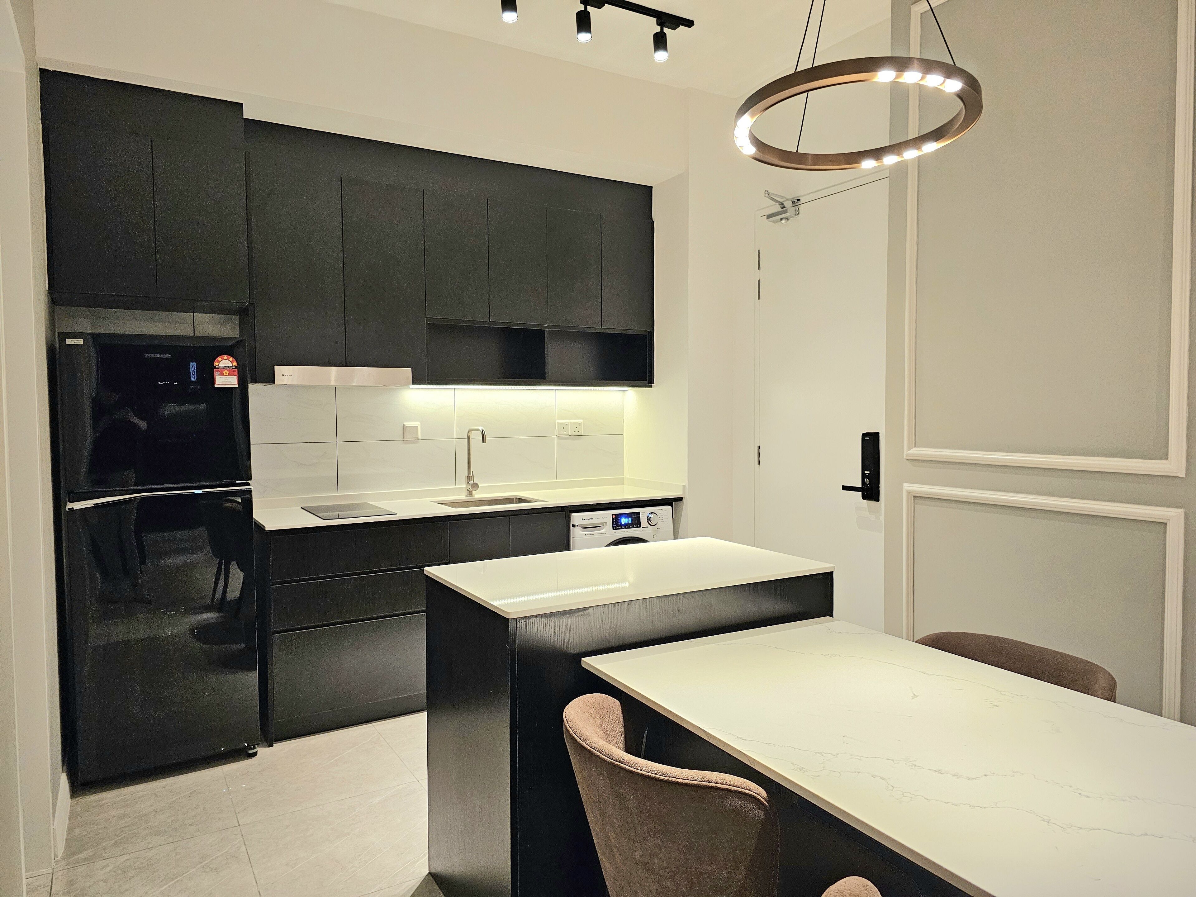 Una cocina moderna con armarios oscuros, encimera blanca y horno empotrado. Hay una zona de comedor con una mesa blanca y dos sillas.