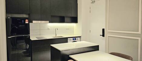 Una cocina moderna con armarios oscuros, encimera blanca y horno empotrado. Hay una zona de comedor con una mesa blanca y dos sillas.