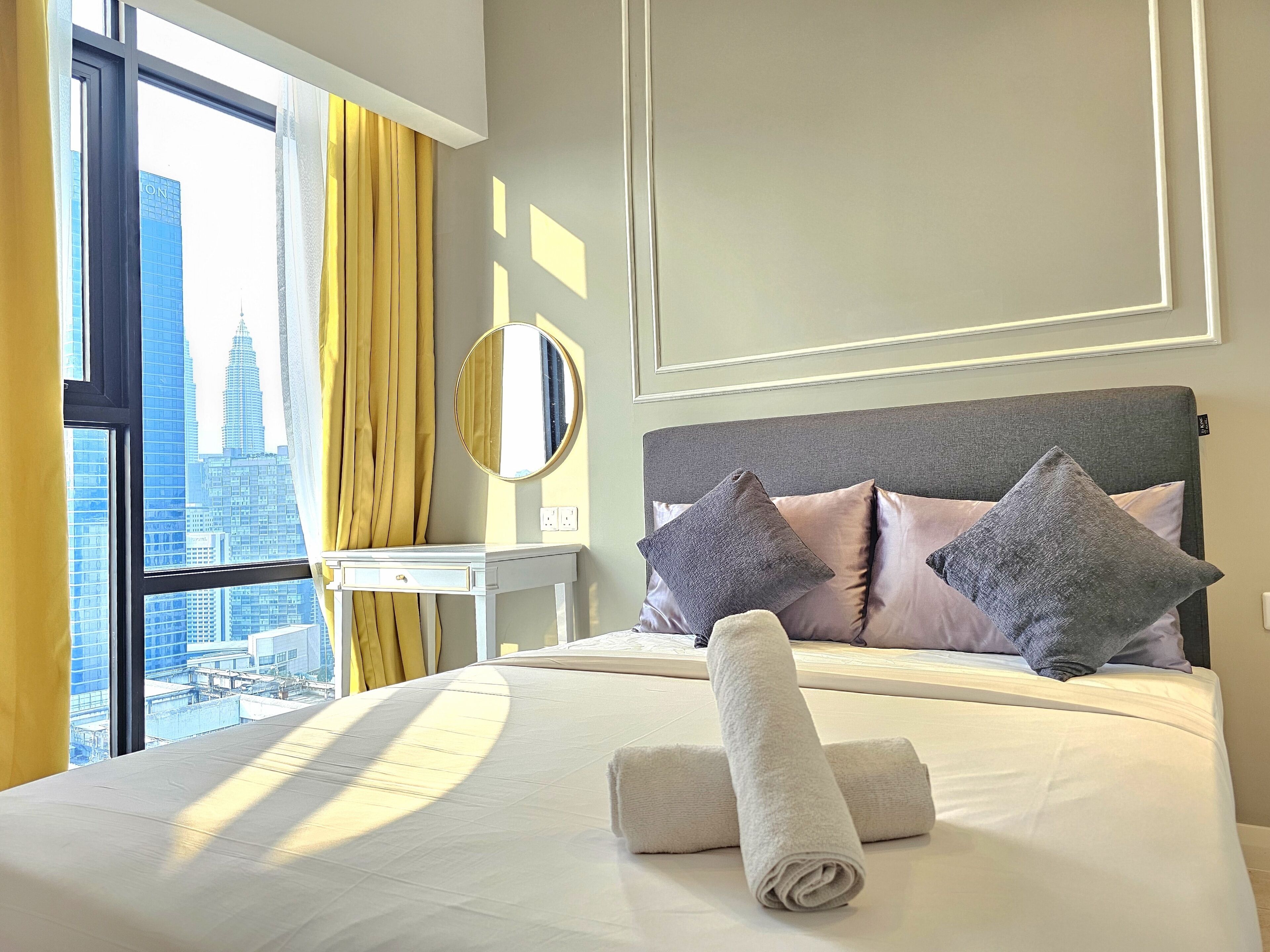 Une chambre d’hôtel avec un grand lit, une table de chevet, un miroir et une vue sur le paysage urbain, visible à travers une fenêtre ornée de rideaux jaunes.