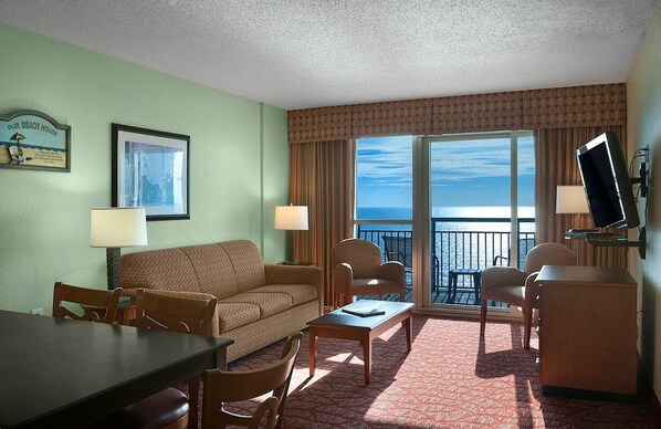 A hotel room with a sofa, chairs, a dining table, a TV, and a balcony with a view of the ocean.