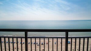A beach view from a balcony with a railing, featuring a sandy beach with umbrellas and a calm sea under a clear sky.