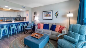 A modern living room with a sofa, ottoman, and bar stools. There is a kitchen area with white cabinets and a blue backsplash. A framed picture hangs on the wall, and a lamp is on a side table.