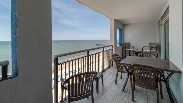 A balcony with a view of the beach, wooden chairs, and a table.