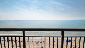 Vista a la playa desde un balcón con barandilla, mostrando una playa arenosa con varias sombrillas y algunas personas.