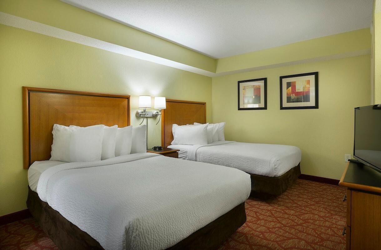 A hotel room with two beds, a nightstand, a lamp, and a television.