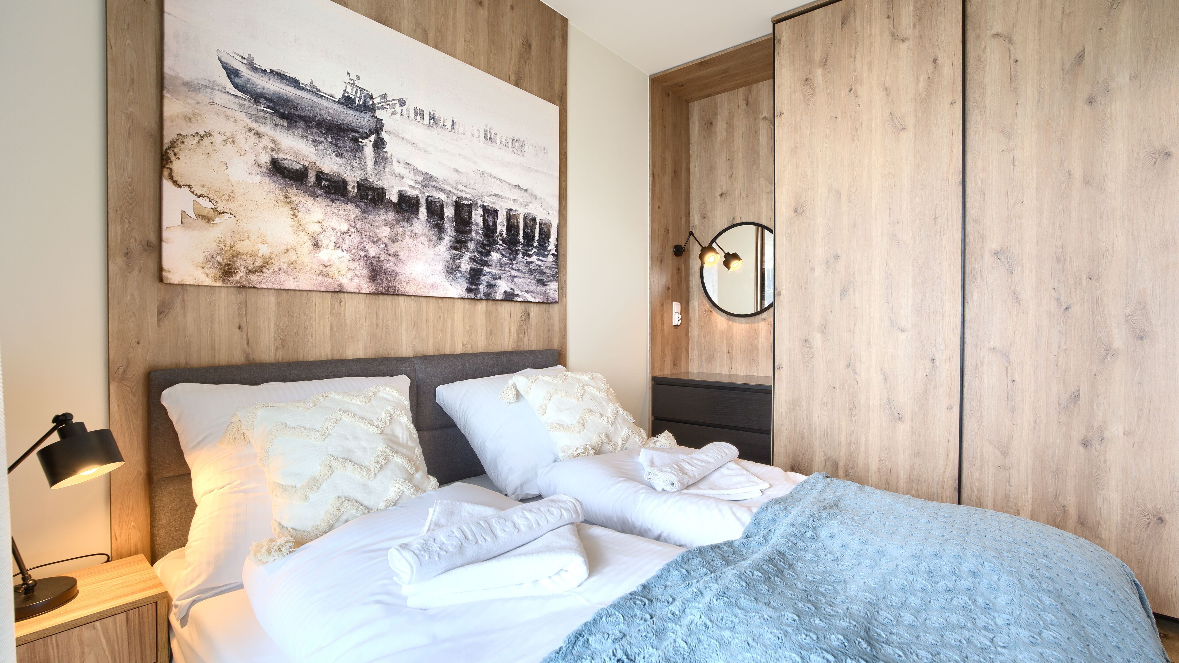 A neatly made bed with white linens and a blue blanket, a wooden headboard, a large abstract painting, a bedside lamp, and a wooden wardrobe.