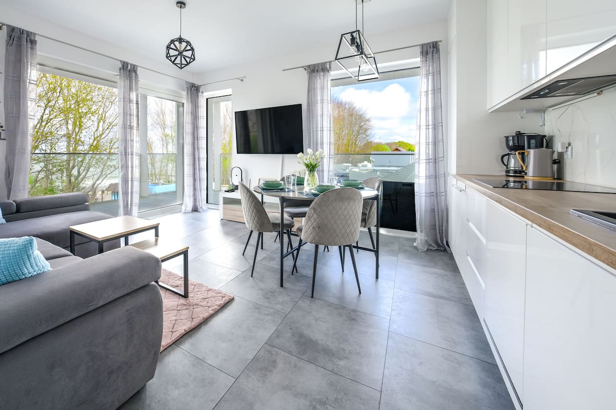 Ein modernes Wohnzimmer mit grauem Sofa, einem Couchtisch und einem Essbereich mit Tisch und Stühlen. Es gibt eine Küchenzeile mit Herd und einem großen Fenster.