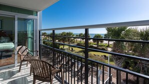 Een balkon met een glazen deur naar een kamer, uitzicht op de oceaan en zitplaatsen met stoelen.