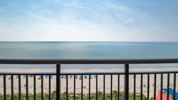 A beach view from a balcony with a railing, a sandy beach, and umbrellas. - Coastal Comfort Retreat with Ocean Views (Myrtle Beach)