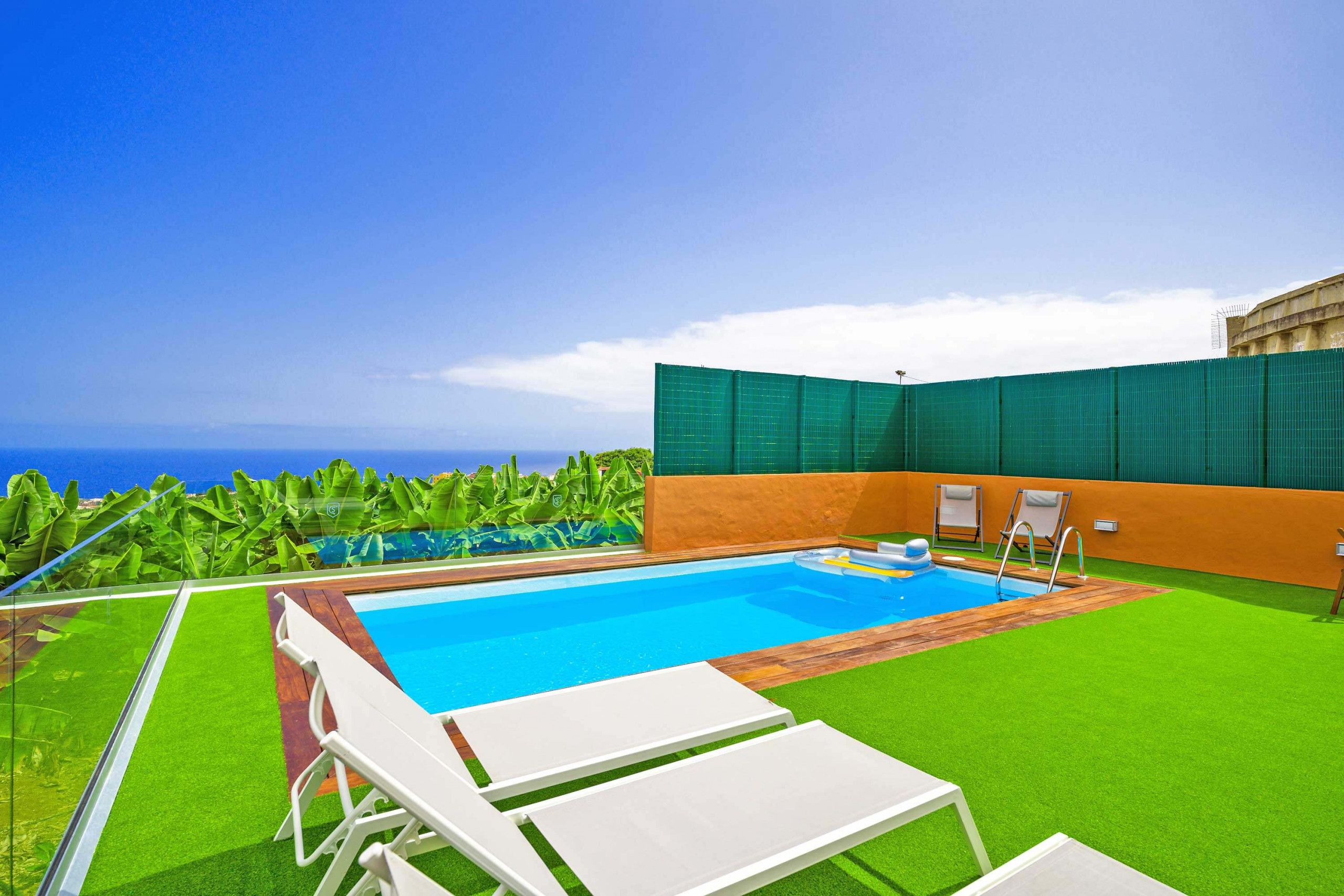 A swimming pool with a view of the ocean, surrounded by green artificial grass and a few lounge chairs.