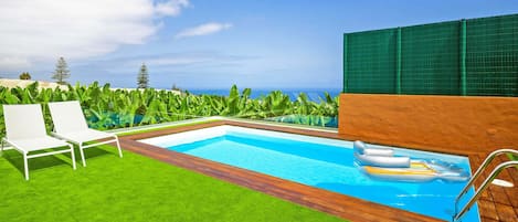 A swimming pool with a wooden deck, surrounded by green grass and white lounge chairs.