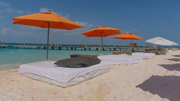 Een strand met rijen witte ligbedden en oranje parasols, een pier op de achtergrond en helderblauw water.