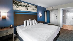 A hotel room with a large bed, a nightstand with a lamp, a painting above the bed, and a door leading to a bathroom. - Wave Watcher's Delight: Oceanfront King Escape (Myrtle Beach)