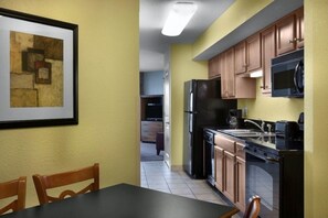 A kitchen with wooden cabinets, a black refrigerator, and a microwave. There is a framed picture on the wall and a dining table with chairs.