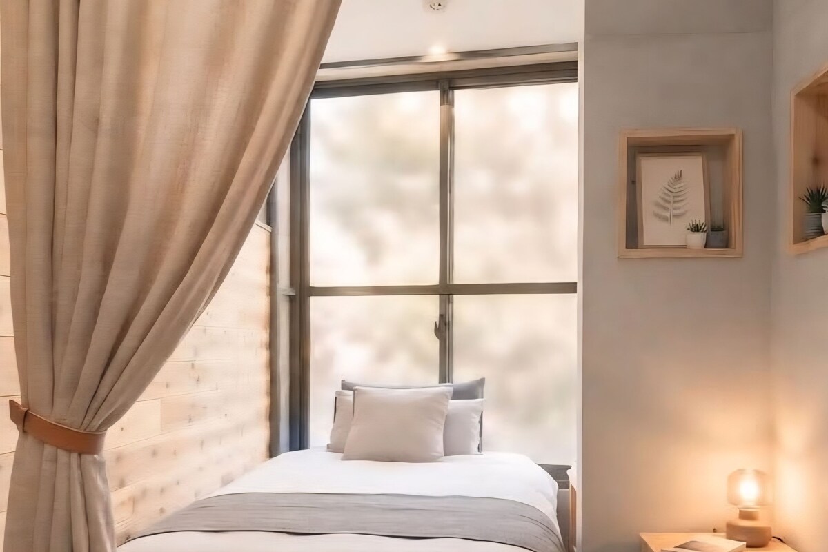 A bedroom with a large window, a bed with white bedding, a bedside table with a lamp, and a wall-mounted plant.